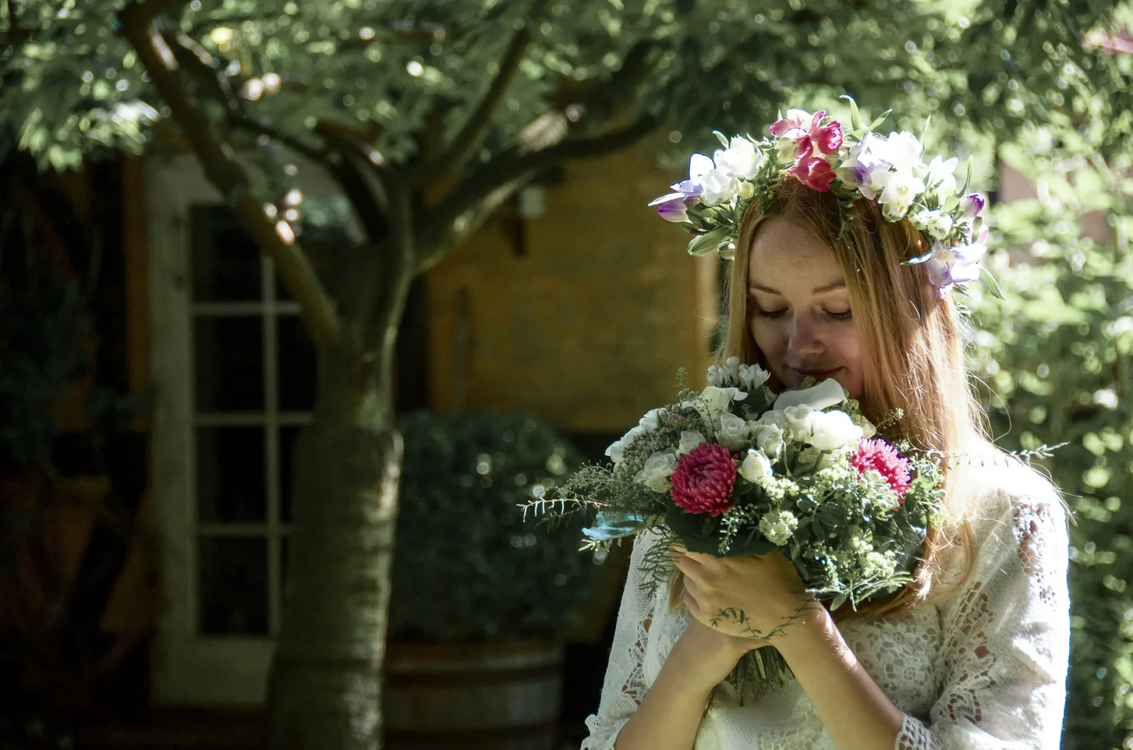 Braut mit Blumenkranz riecht im Schatten eines Baumes an ihrem rosa-weißen Brautstrauß