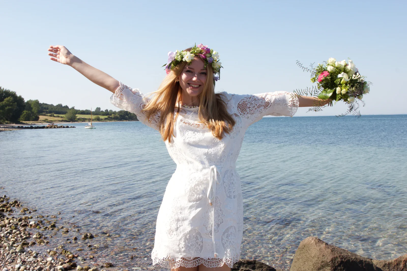 Lachende Braut im weißen Spitzenkleid mit Blumenkranz und Brautstrauß am steinigen Meeresufer