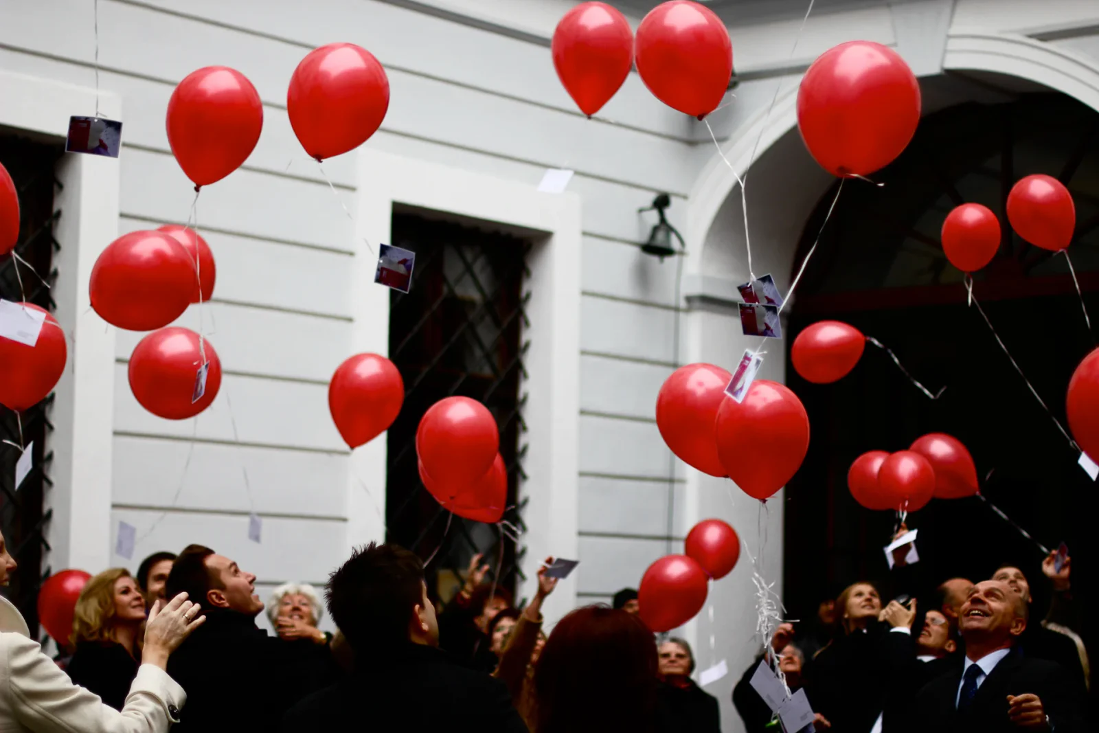 Hochzeitsgäste lassen vor einem hellen Gebäude rote Luftballons mit Karten steigen
