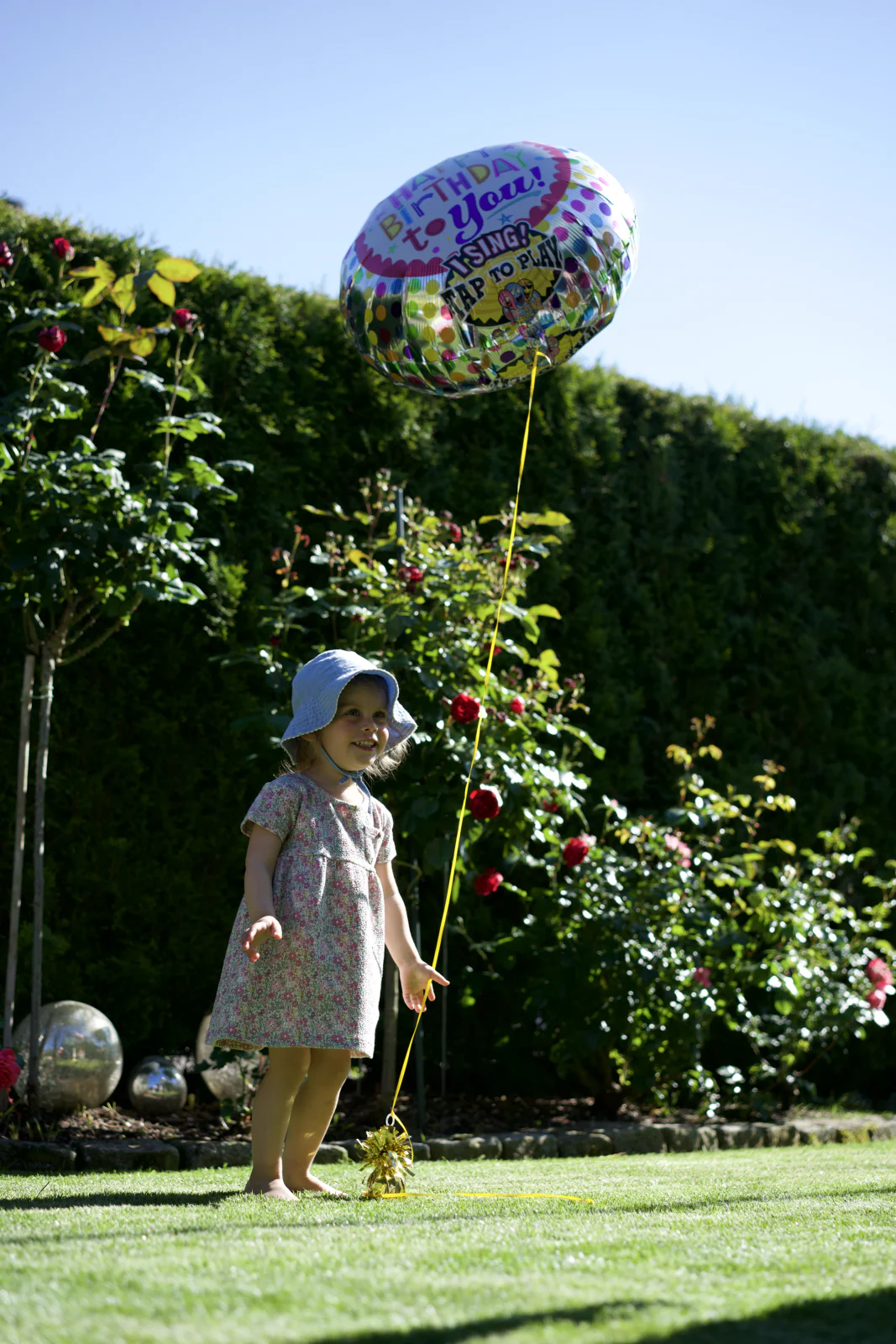 Kleines Kind mit Sonnenhut steht barfuß im Garten neben einem bunten Geburtstagsballon