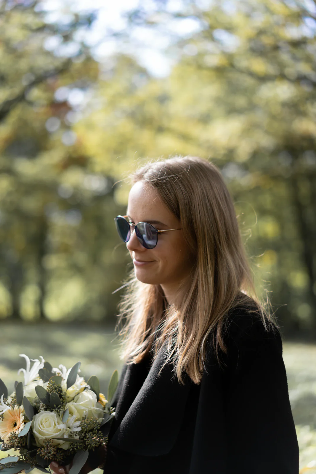 Frau mit Sonnenbrille und schwarzem Mantel hält einen hellen Blumenstrauß im sonnigen Park