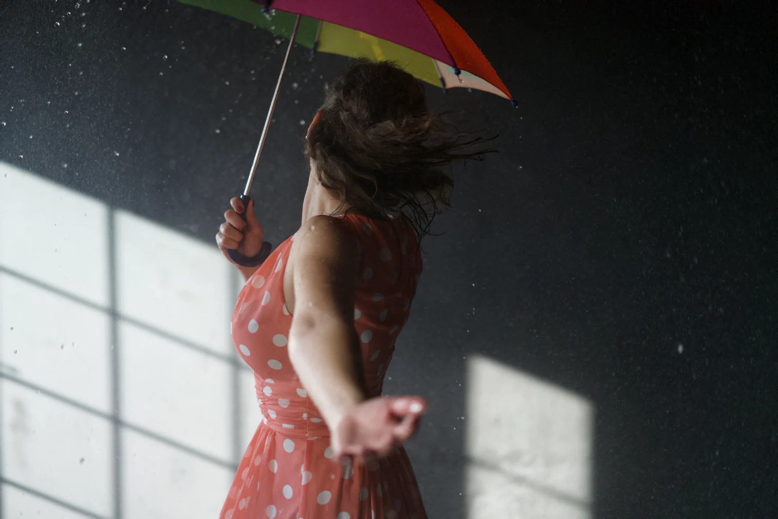 Frau im gepunkteten Kleid dreht sich mit buntem Regenschirm durch Wassertropfen im Studio