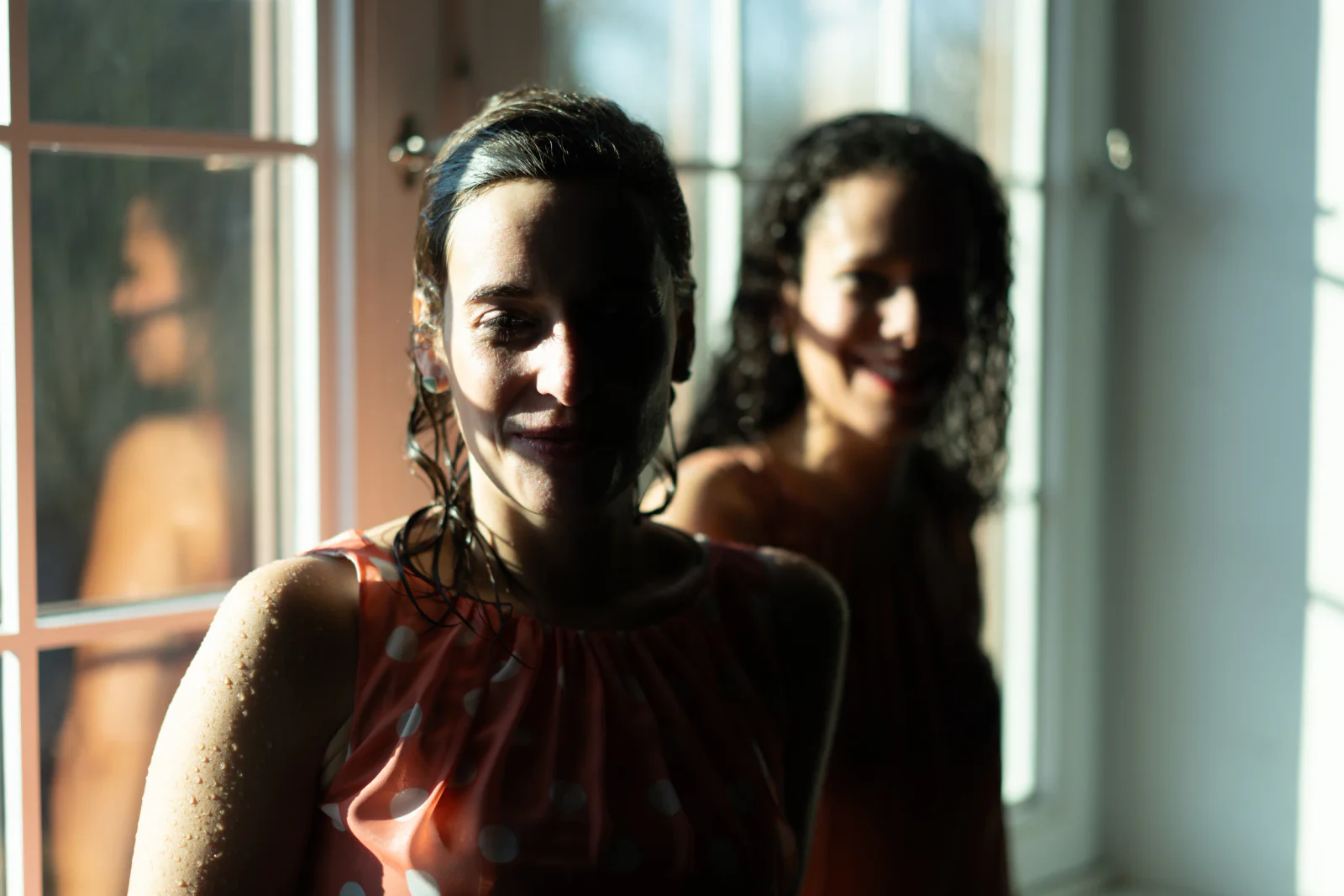 Zwei Frauen mit nassem Haar im harten Fensterlicht, die vordere lächelnd im Fokus