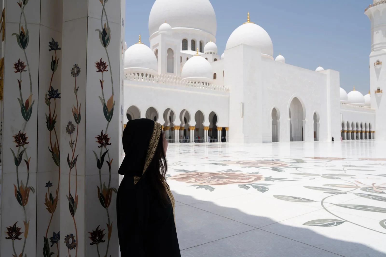 Frau in schwarzer Kleidung blickt aus einem verzierten Säulengang auf eine weiße Moschee